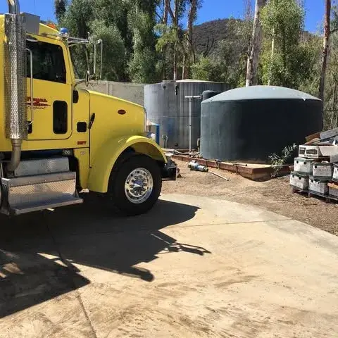 Side view of a yellow semi truck parked in front of a water tank in the middle of the day.